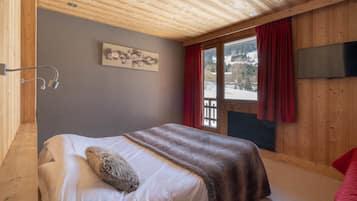 Un dormitorio con techo de madera, cama con colchón blanco y manta marrón, televisor montado y ventana con cortinas rojas.