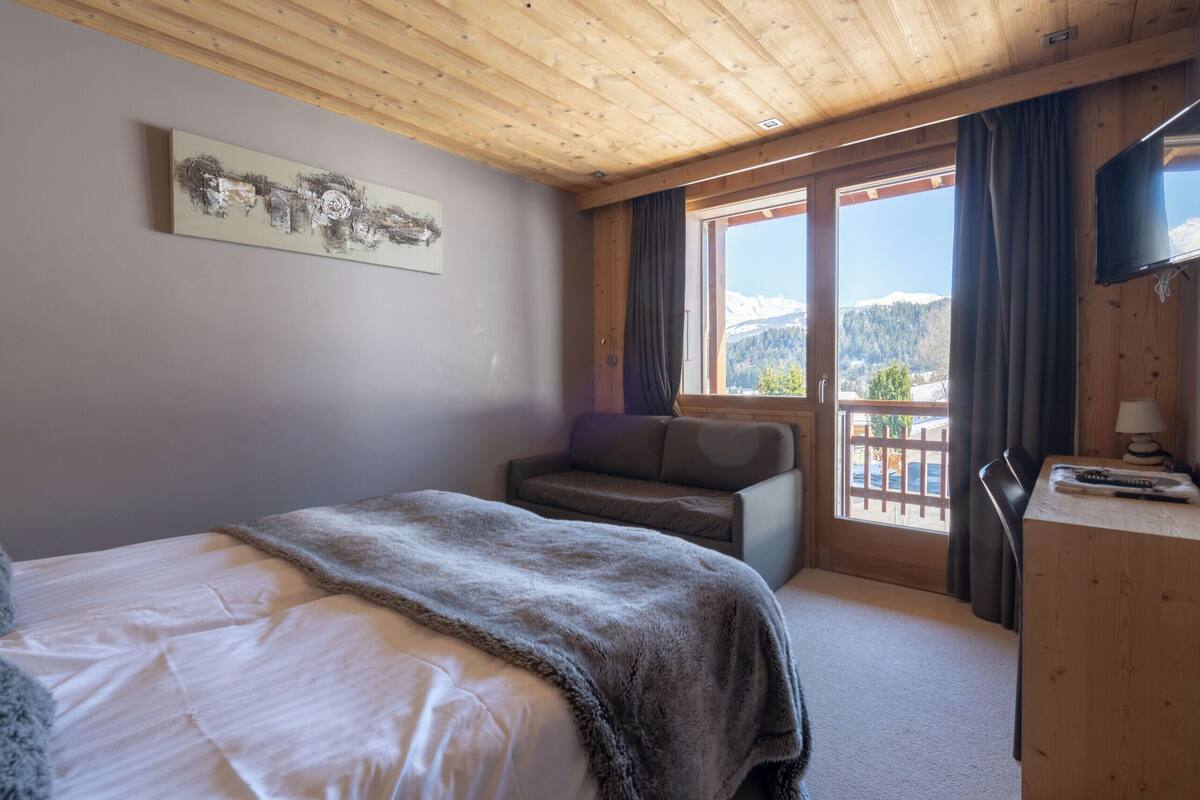 A room with a bed, a sofa, a desk, and a TV. There is a window with a view of mountains.