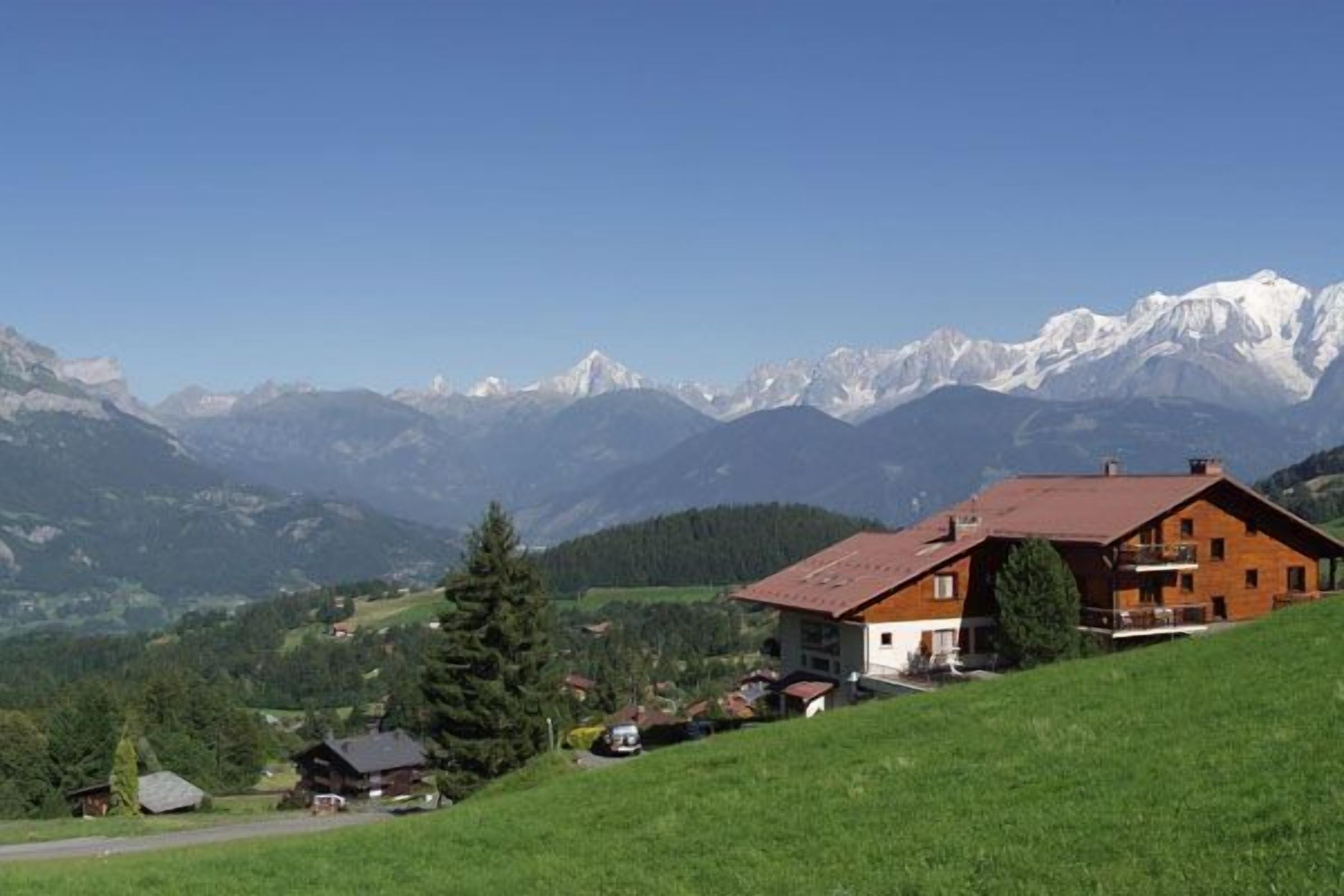 A wooden house with a sloped roof on a hillside, surrounded by green fields and mountains in the background.