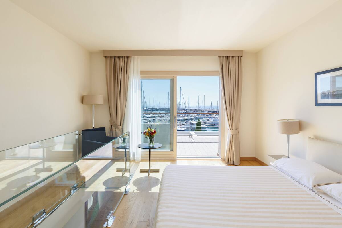A hotel room with a bed, a glass table, a chair, a small table with a vase, and a view of boats through the window.