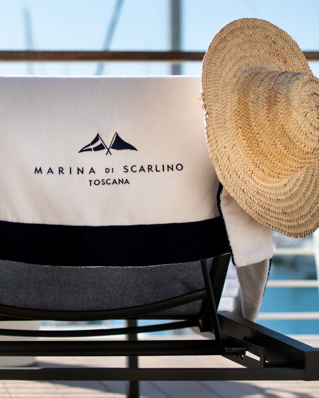 A lounge chair with a towel and a straw hat, branded with 'MARINA DI SCARLINO TOSCANA' logo.