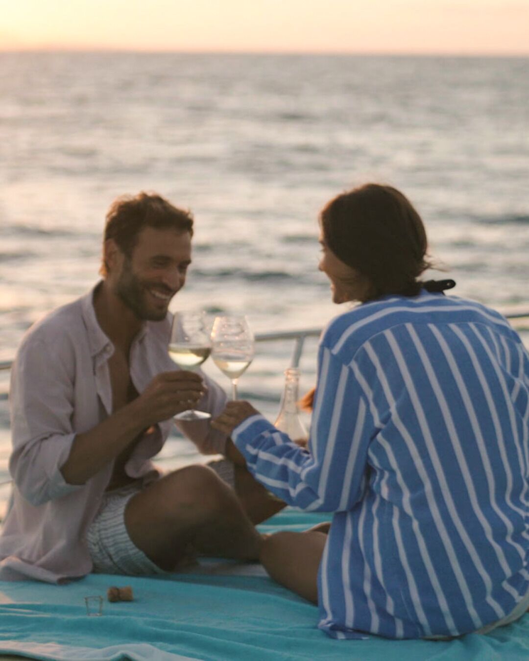 Deux personnes sont assises sur un tapis bleu, près de l’eau, et tiennent chacune un verre de vin.
