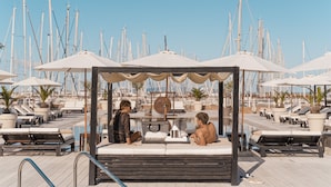Un área junto a la piscina con sillones de descanso y sombrillas, una terraza de madera y un fondo de veleros.