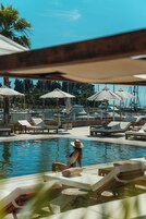 Un área de piscina con sillones de descanso y sombrillas, rodeada por un puerto deportivo con barcos.
