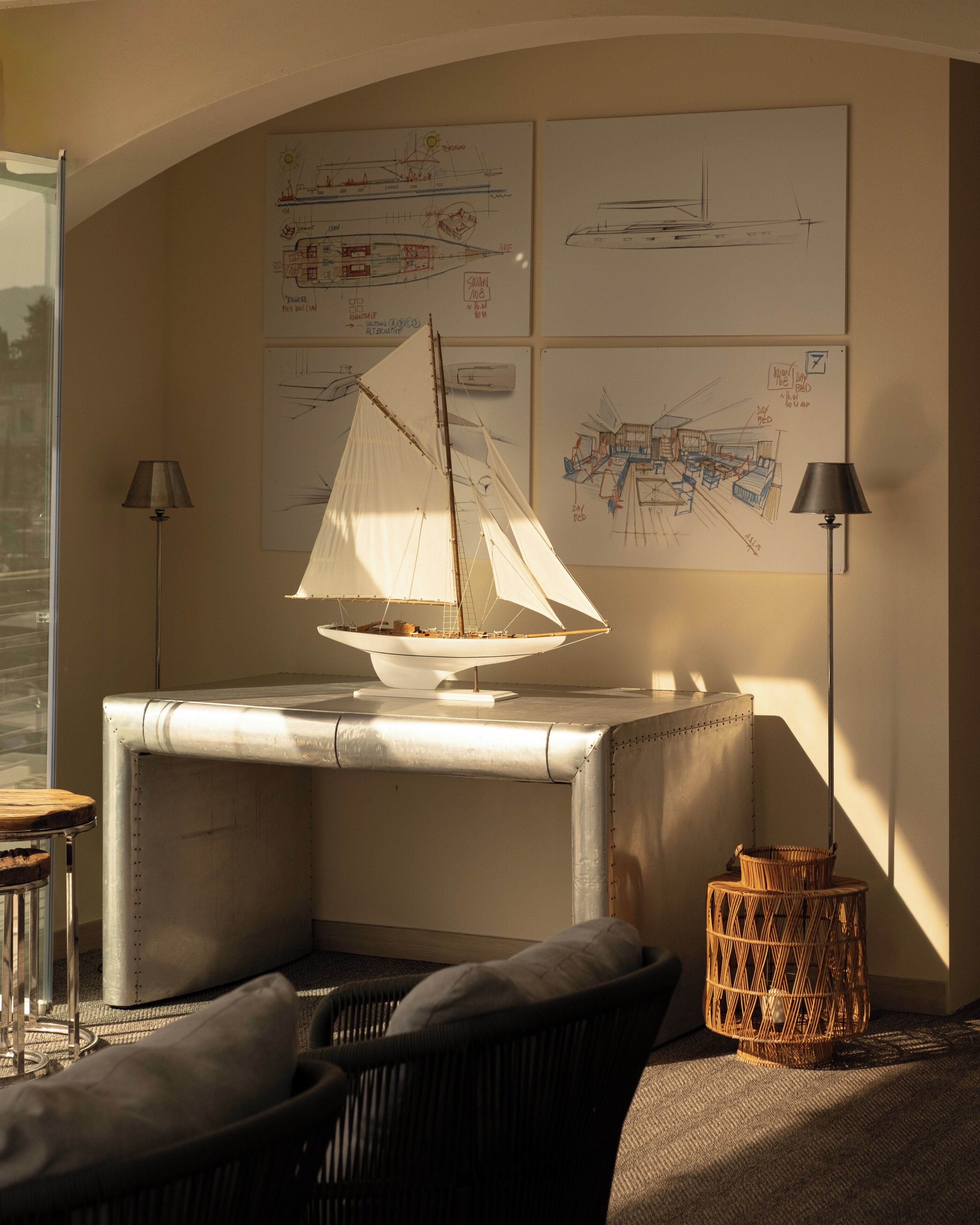 Une maquette de voilier est posée sur une table, entourée d’un coin salon confortable avec une lampe, un panier en osier et des plans d’architecture accrochés au mur.