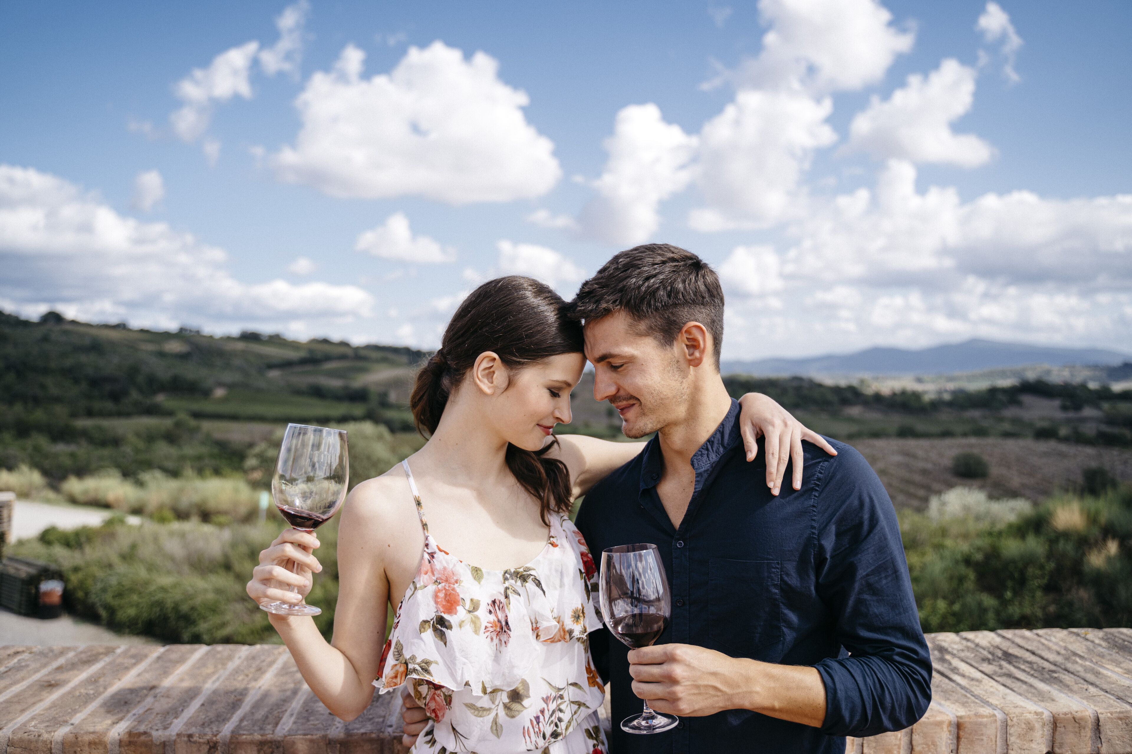 Un couple, chacun tenant un verre de vin, contemple un vignoble sous un ciel dégagé.