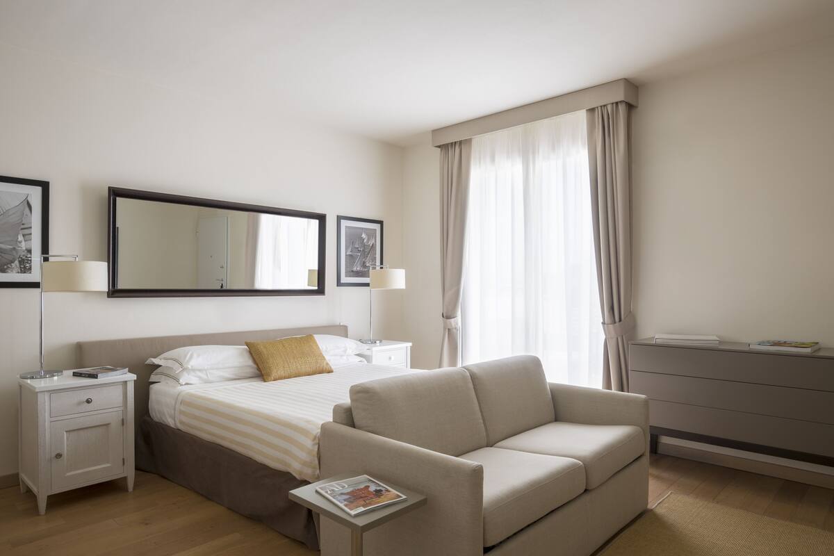 A hotel room with a bed, a sofa, a nightstand, a dresser, and a large window with curtains.