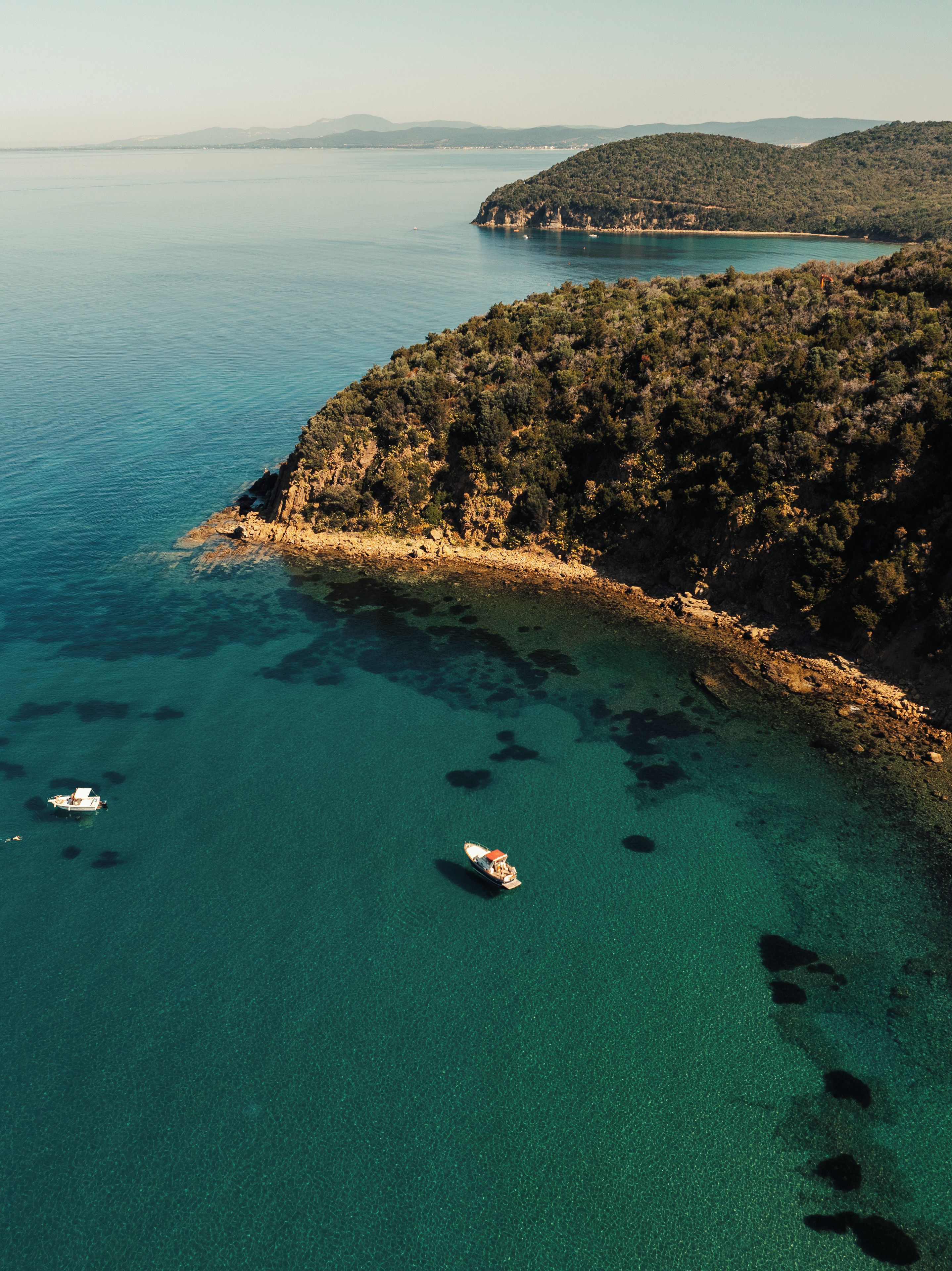 An aerial view of a coastal area with clear turquoise waters, rocky shoreline, and boats.