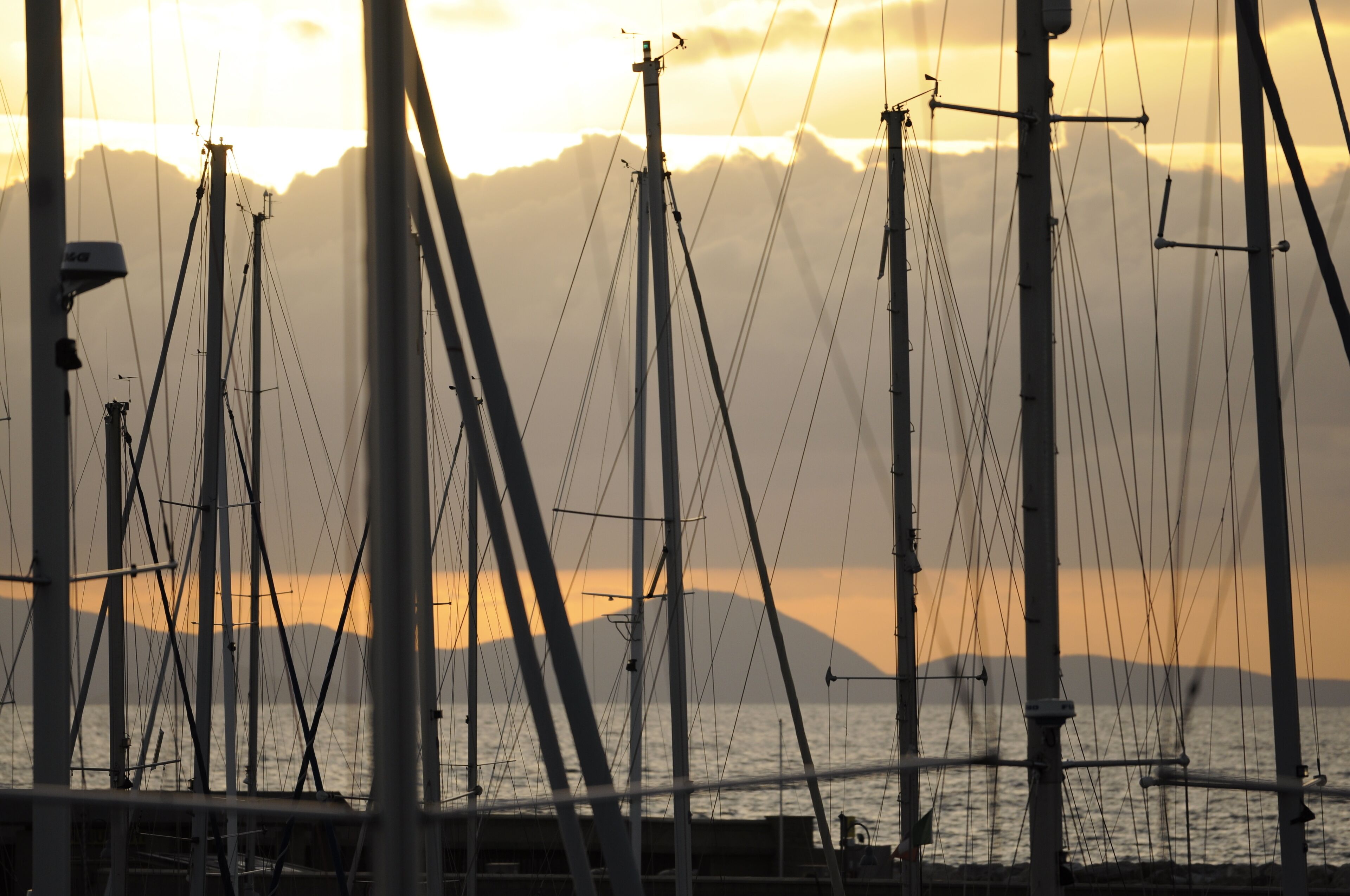 Een jachthaven met meerdere zeilboten afgemeerd, een brug op de achtergrond en een zonsondergang boven het water.