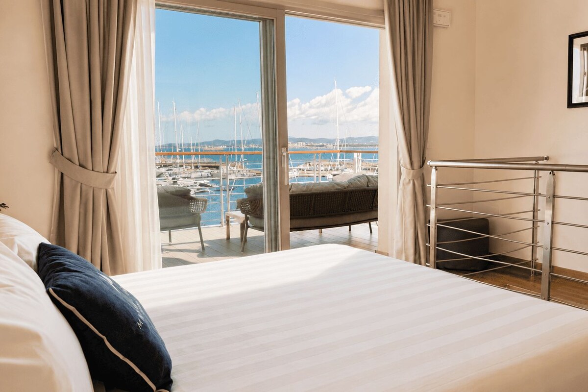 A hotel room with a bed, a view of a marina, and a balcony with a railing.