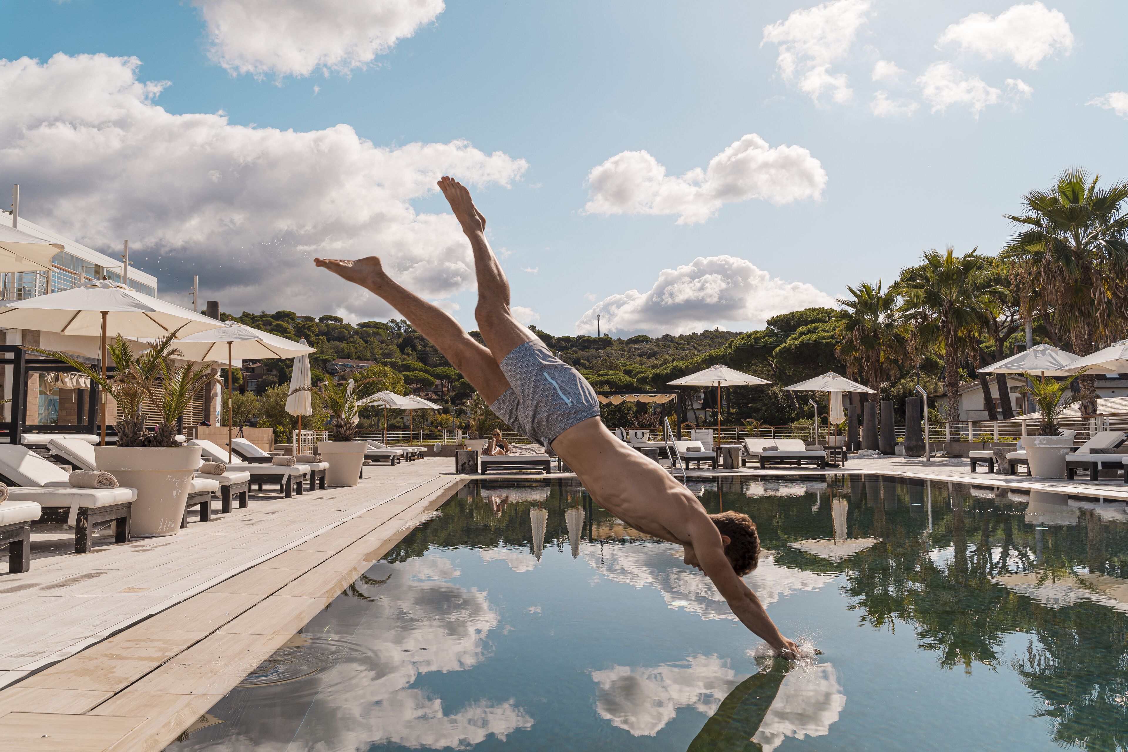 A man diving into a pool with lounge chairs and umbrellas in the background.