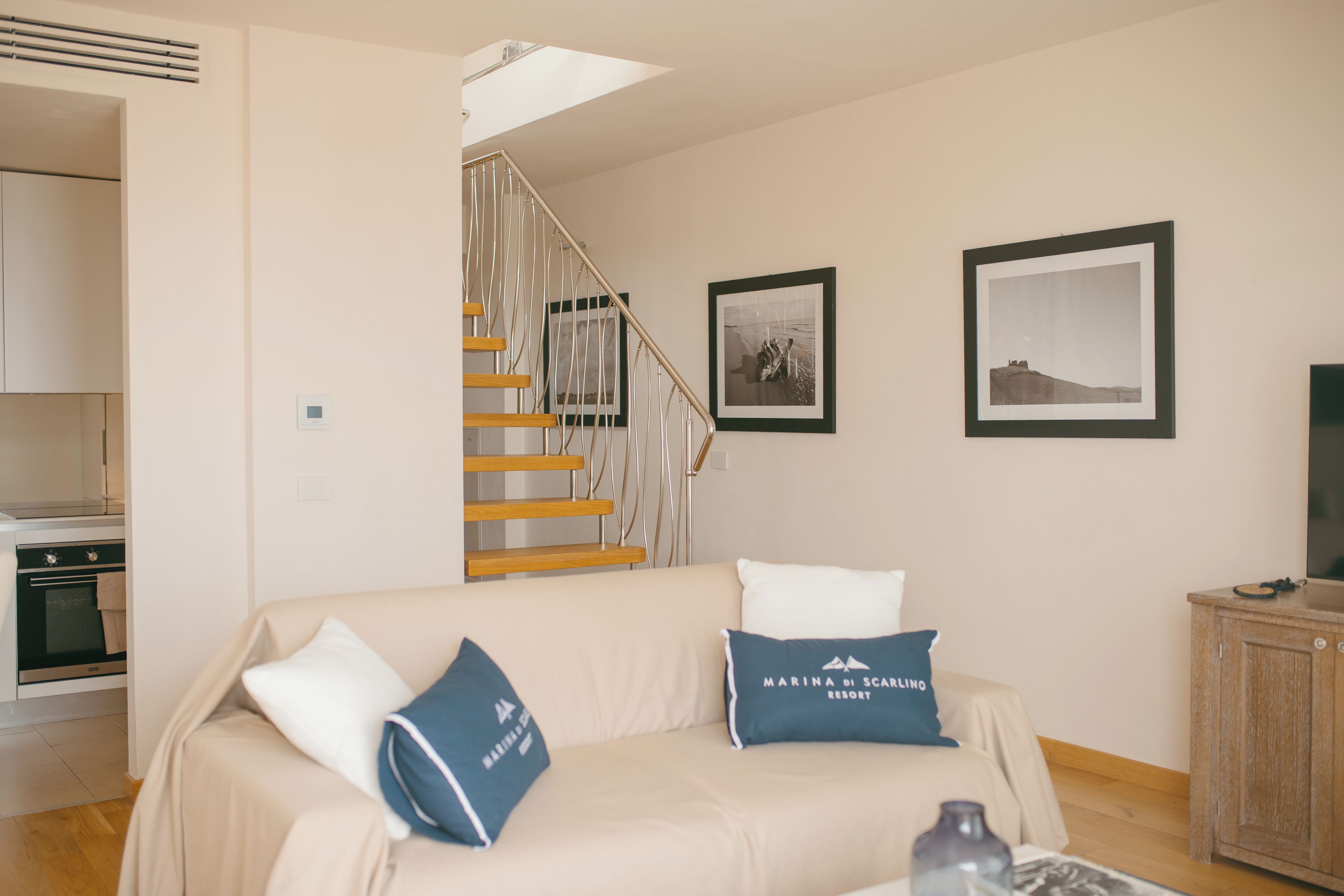 Un salon avec un canapé beige, deux coussins, un escalier en bois et deux tableaux encadrés accrochés au mur.