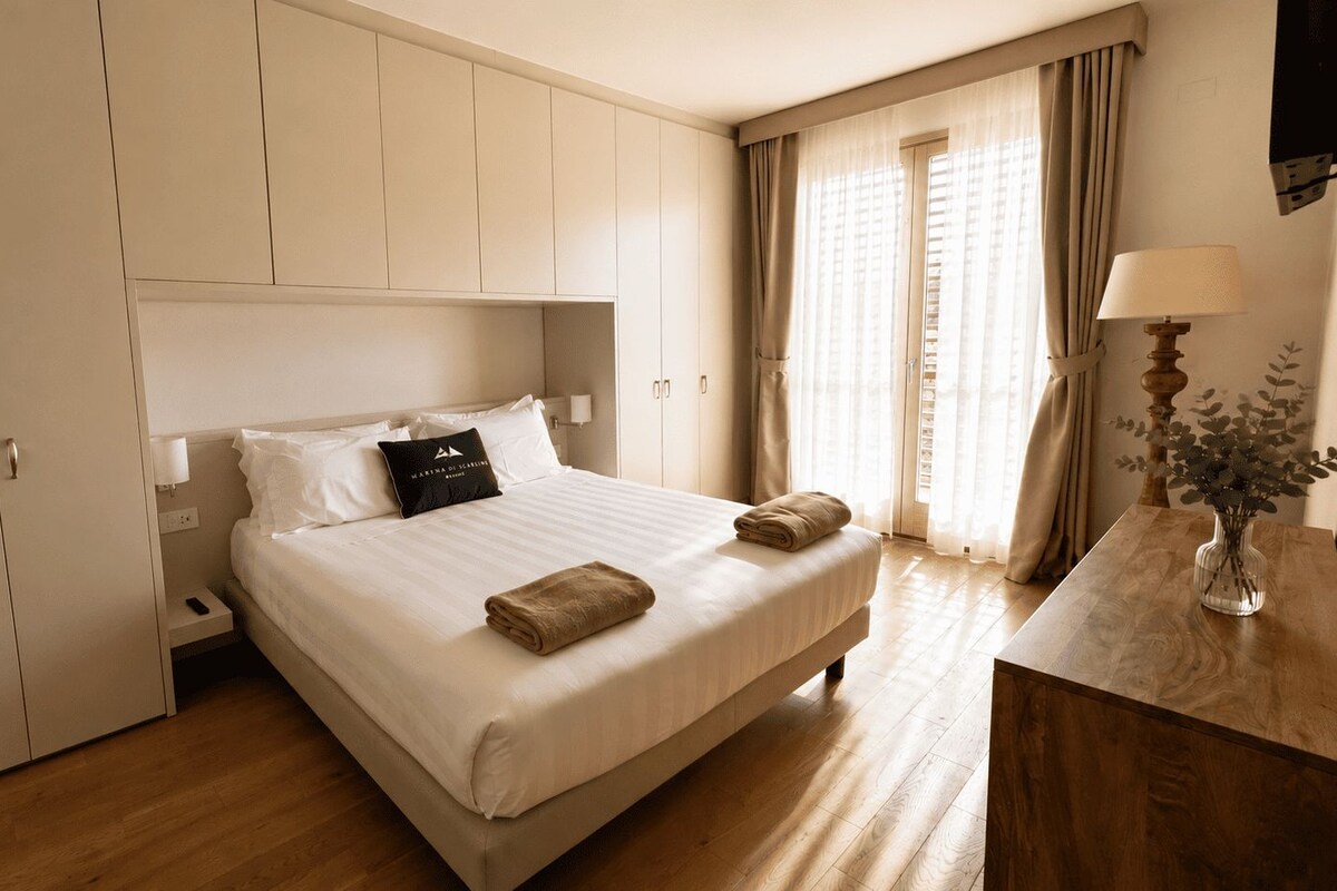 A hotel room with a bed, bedside table, lamp, and a view of the outside.