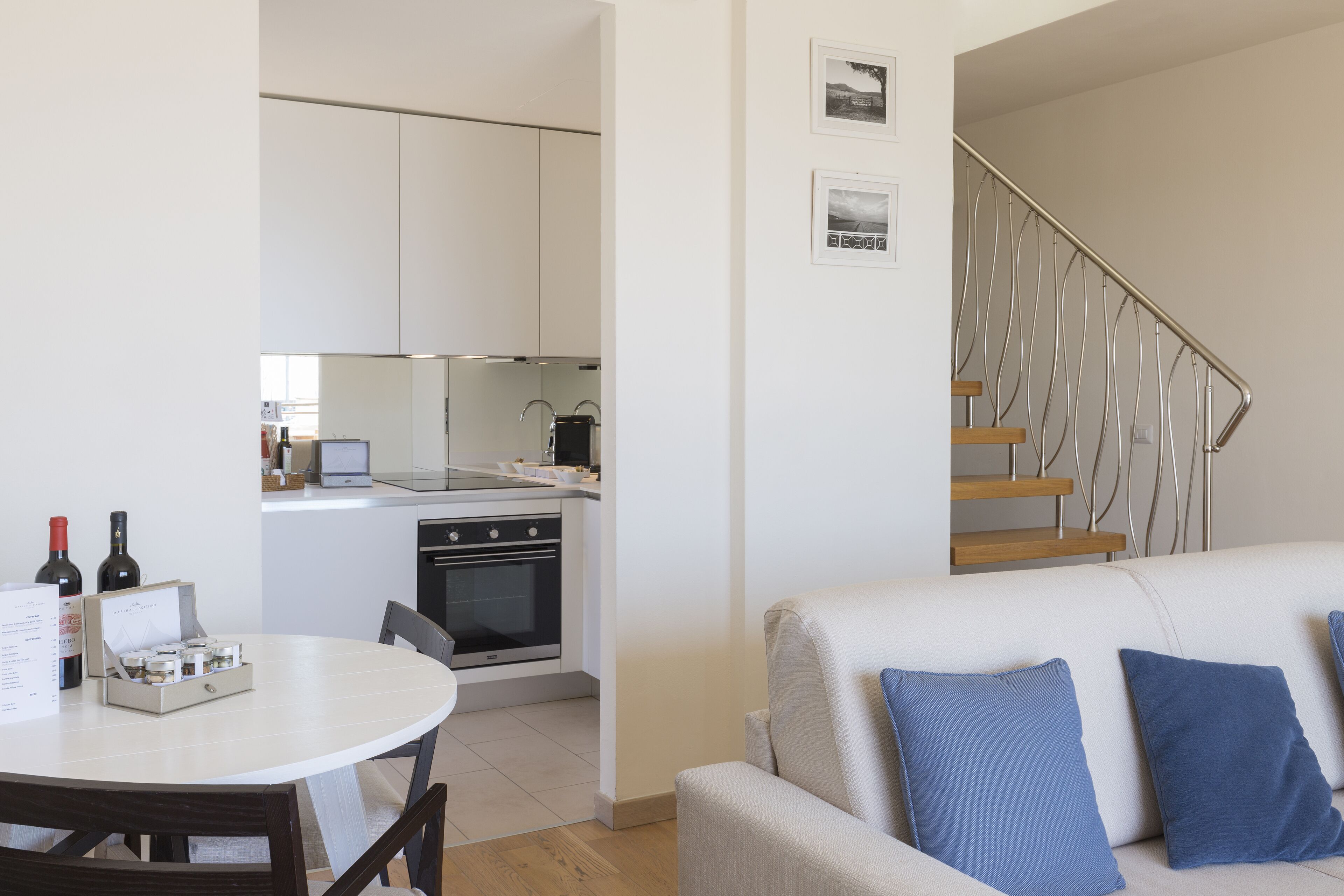 A modern kitchen with white cabinets, a dining table with chairs, a sofa with blue cushions, and a staircase with metal railings.