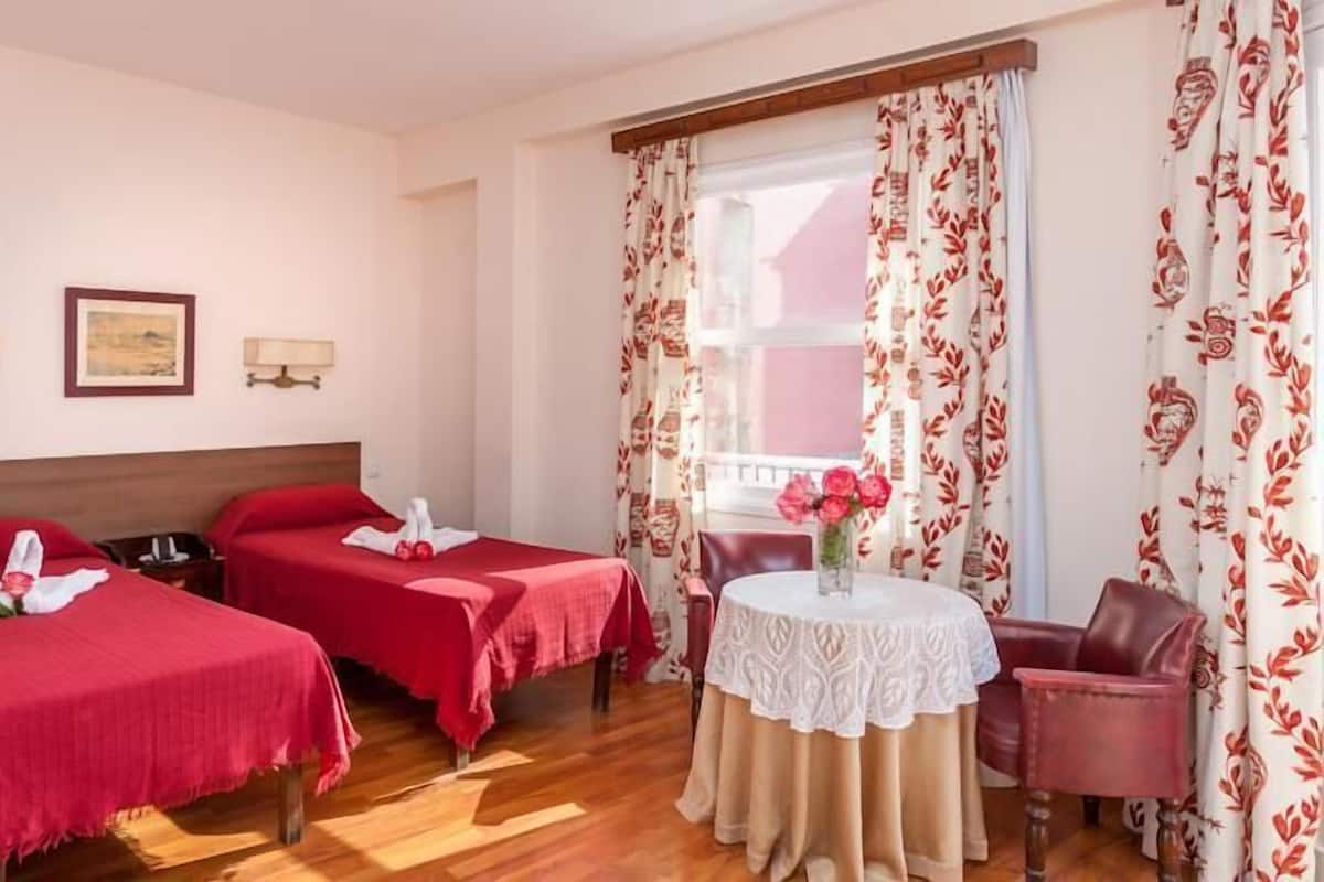 Een hotelkamer met twee eenpersoonsbedden, een klein rond tafeltje met een bloemenvaas, en een raam met bloemen gordijnen.