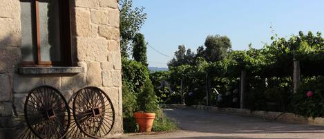 A stone building with a window and two large wheels, a potted plant, and a garden with trees and flowers.