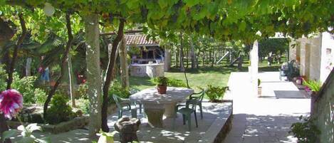 Un patio con una mesa y sillas de piedra, rodeado de plantas en macetas y una pérgola cubierta de hiedra.