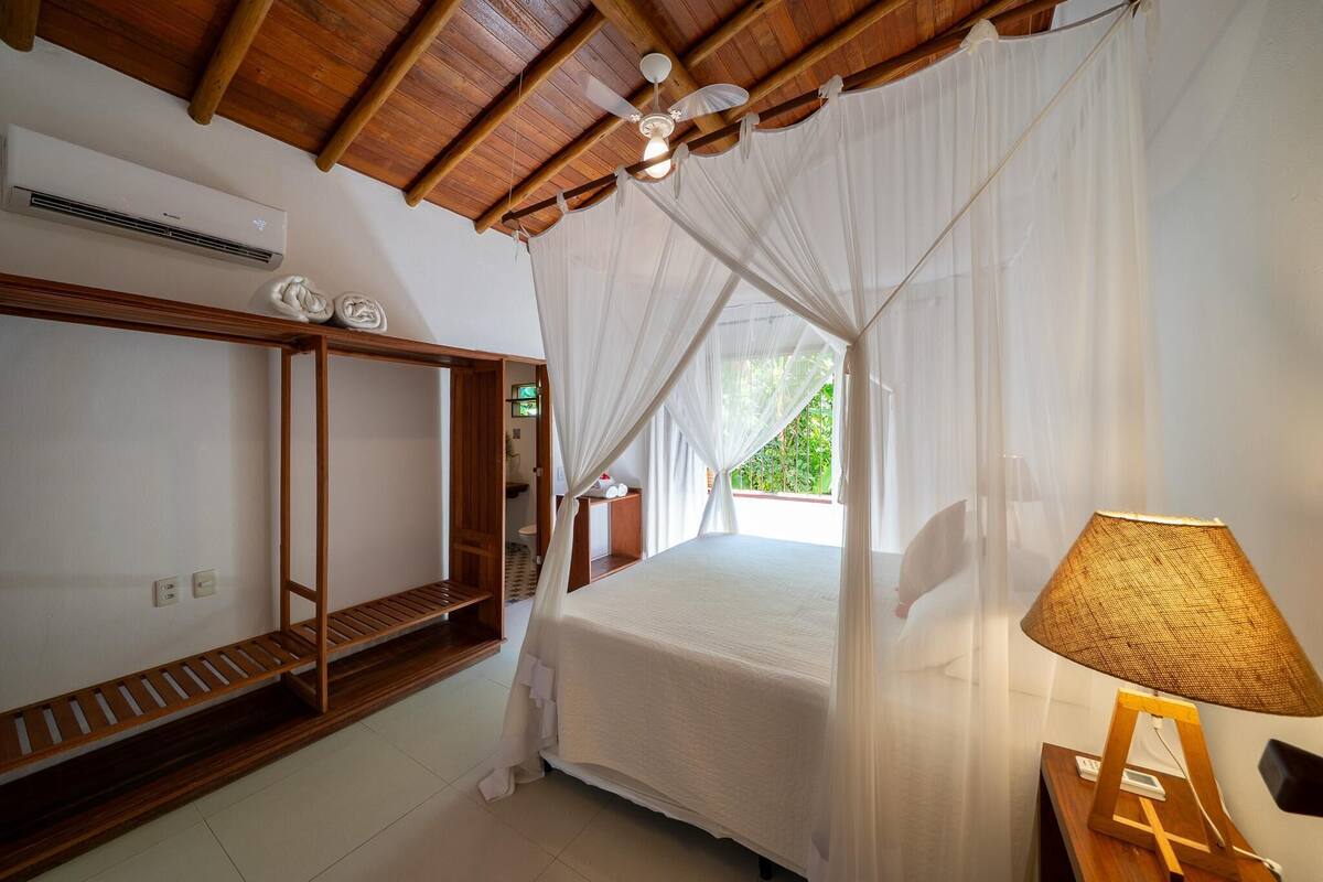 Quarto com teto de madeira, cama com cortinas brancas, mesa de cabeceira com abajur e prateleira com objetos decorativos.