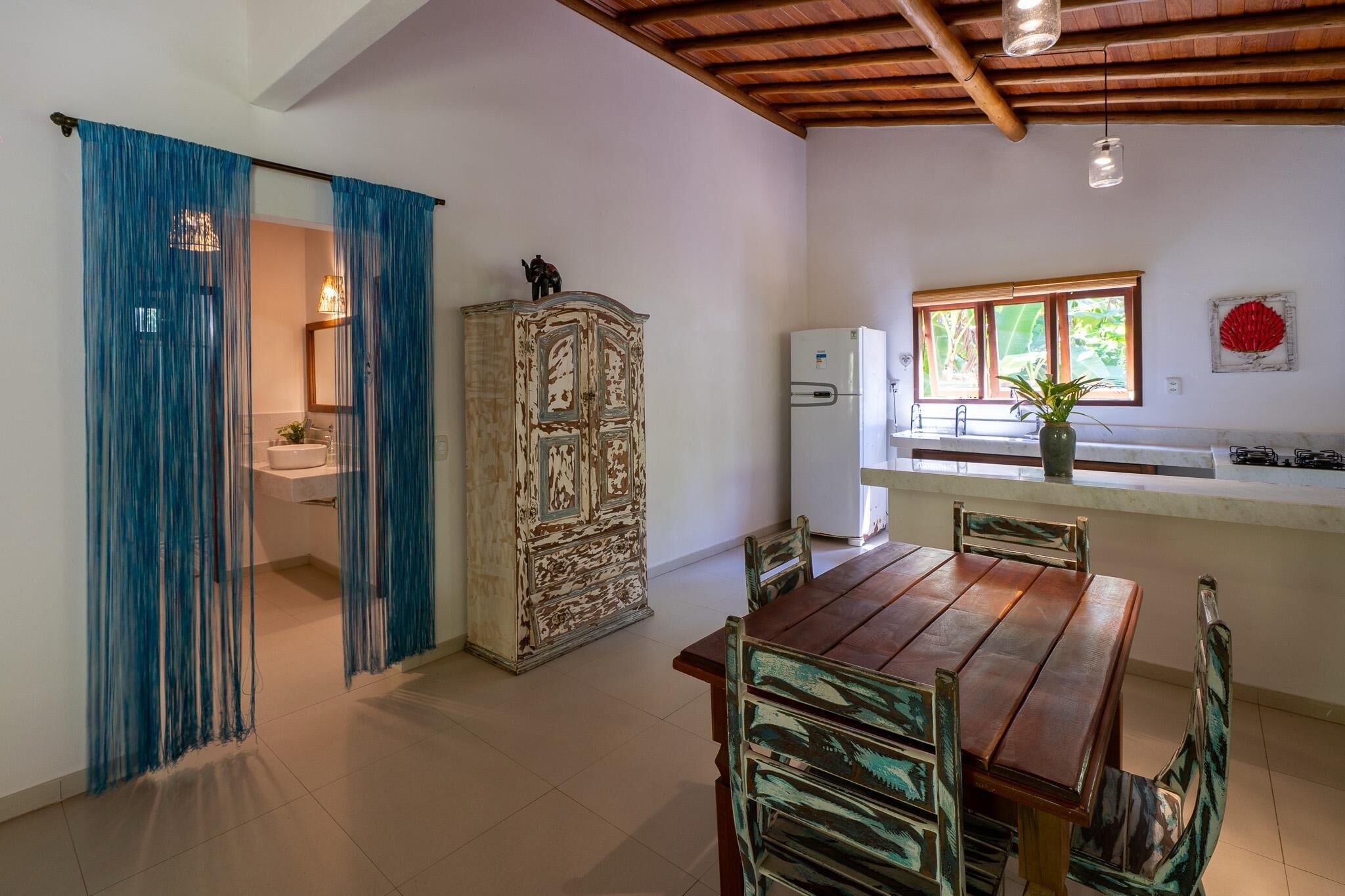 A kitchen with a wooden table and chairs, a refrigerator, and a window with a view of greenery.