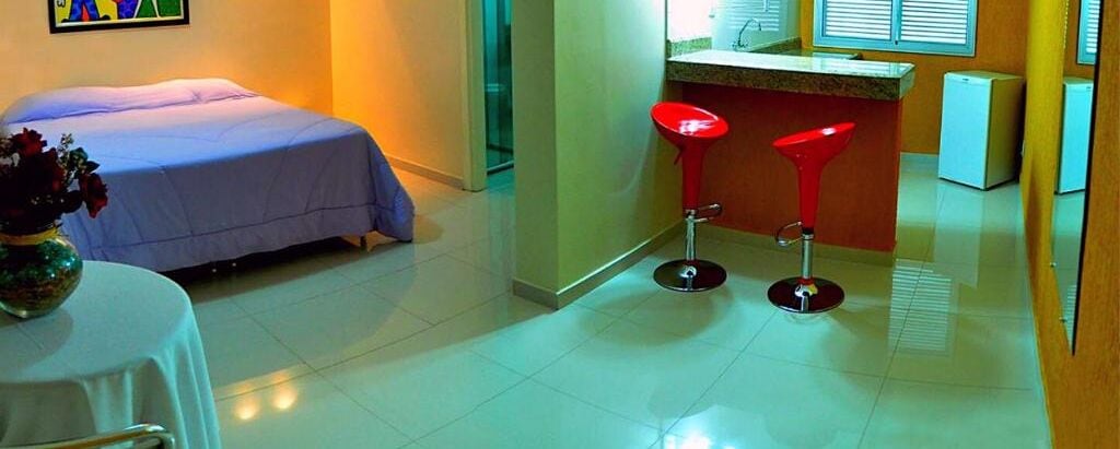Una habitación de hotel con una cama, una mesita con un florero y dos taburetes rojos.