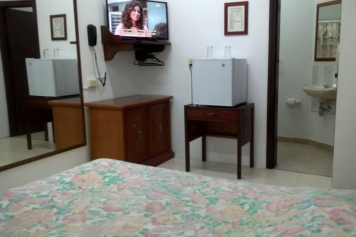 A bedroom with a bed, a wooden wardrobe, a small table, a television, and a bathroom with a sink and mirror.