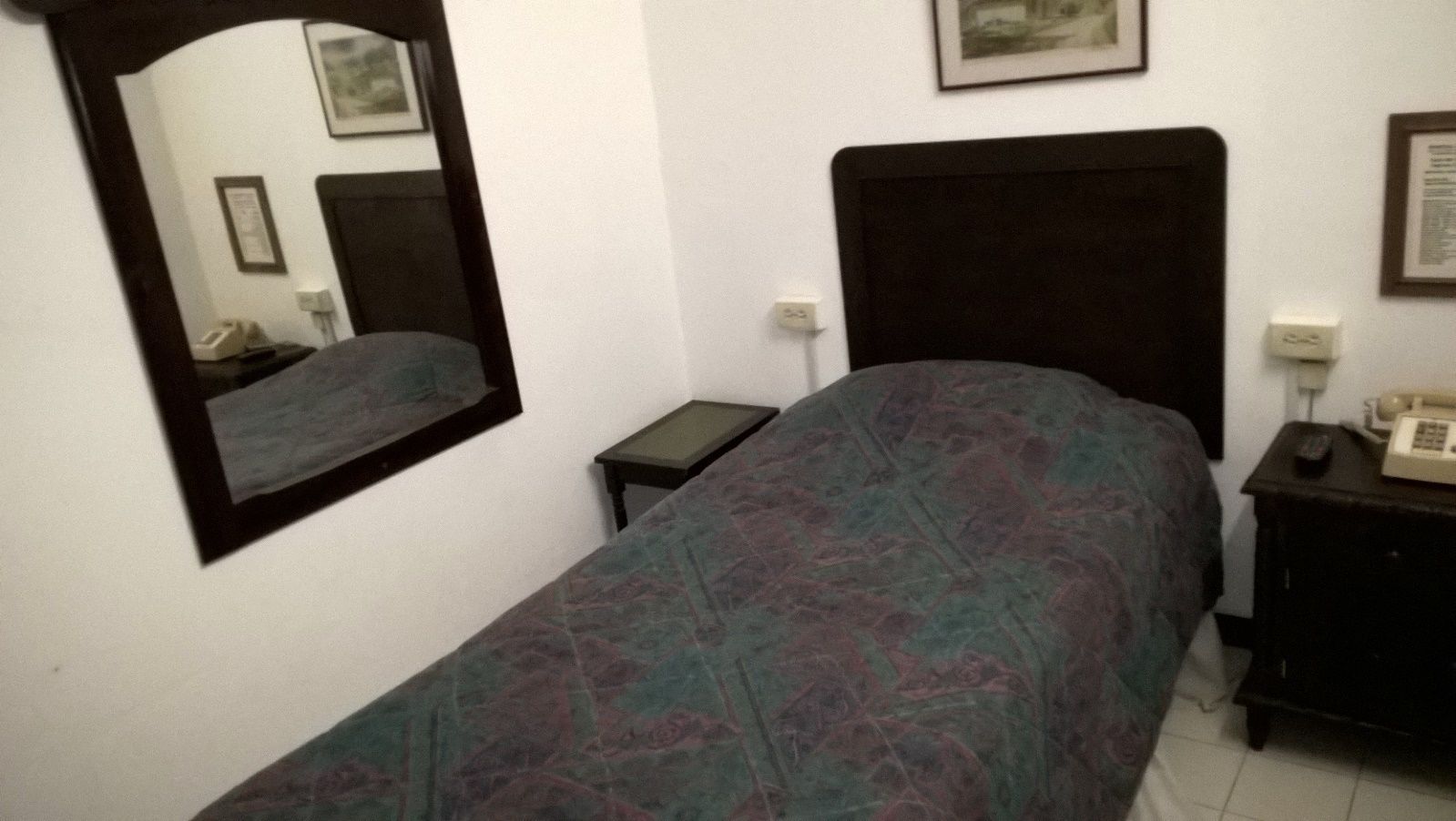 A bedroom with a bed, a nightstand, a bedside table, a mirror, and a wall-mounted telephone.