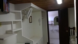 A hotel room with a wooden ceiling, a white wall with a mounted fan, a white shelf unit, a door leading to a bathroom, and a small framed picture. - Hostal San Fernando (Cali)
