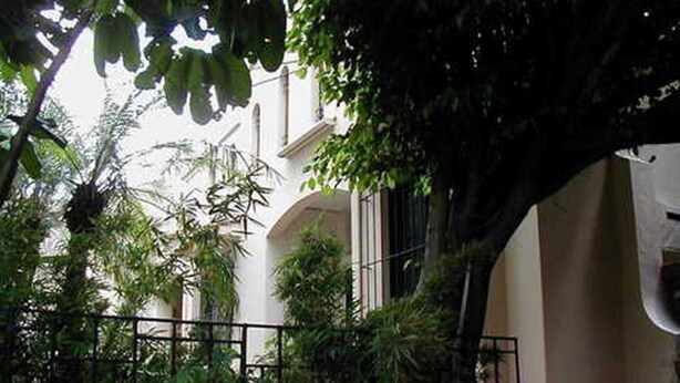 A courtyard with a metal fence, surrounded by lush greenery and a building with a white facade and arched windows. - Hostal San Fernando (Cali)
