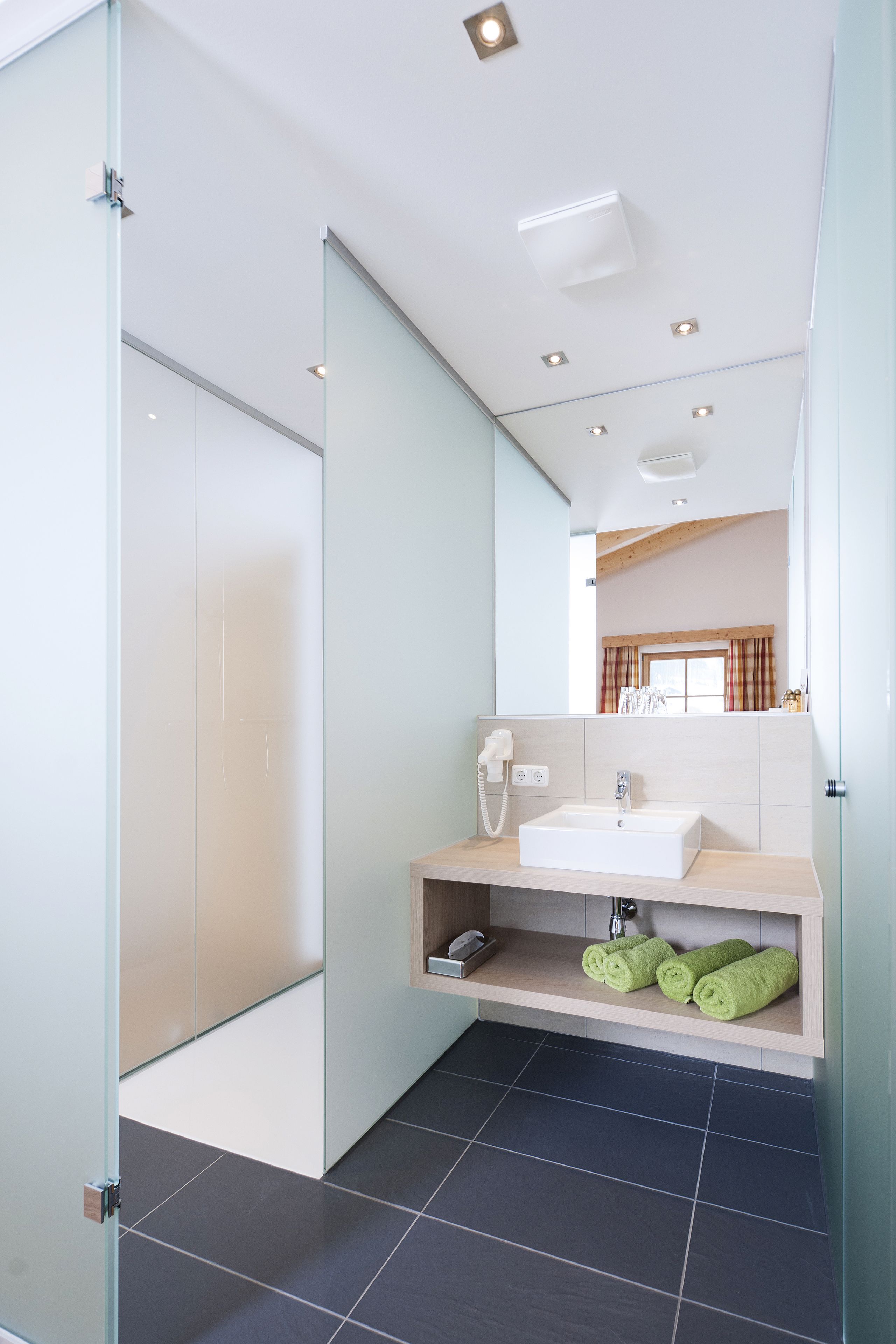A modern bathroom with a glass shower enclosure, a white sink, and a mirror reflecting a window with curtains.