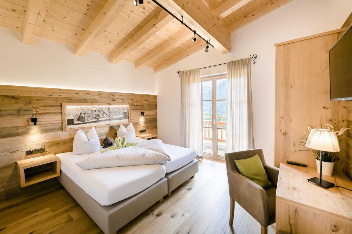 A modern bedroom with a wooden ceiling, a large bed with white bedding, a desk with a lamp, and a view of the outdoors through a window.