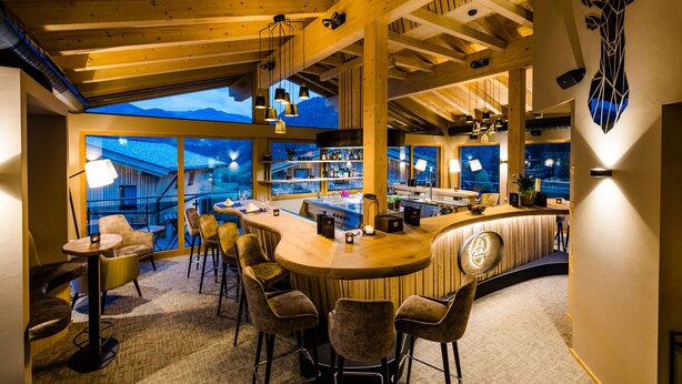 A bar area with a curved counter, multiple bar stools, and a view of the mountains through large windows. - Beim Hochfilzer (Soell)