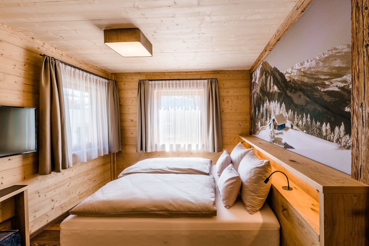 A wooden cabin room with a bed, curtains, a TV, and a mountain painting.