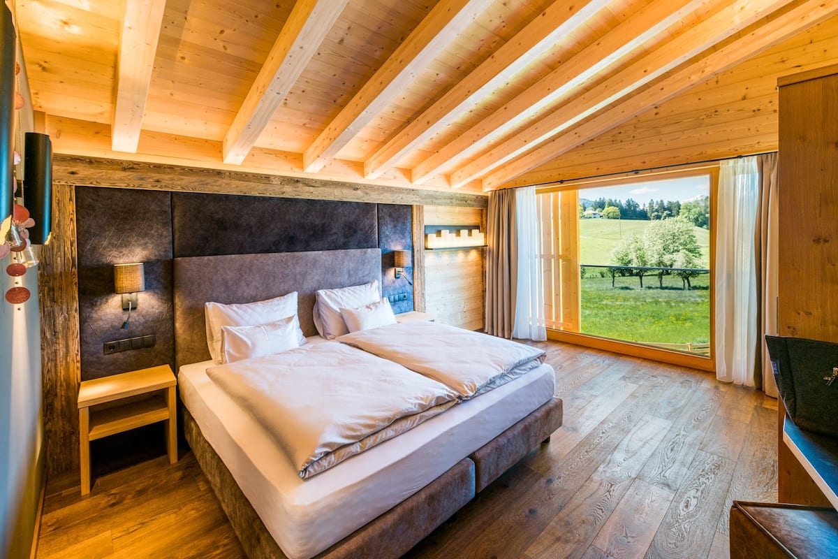 A bedroom with a large bed, wooden floors, a headboard, bedside lamps, and a view of a green landscape.