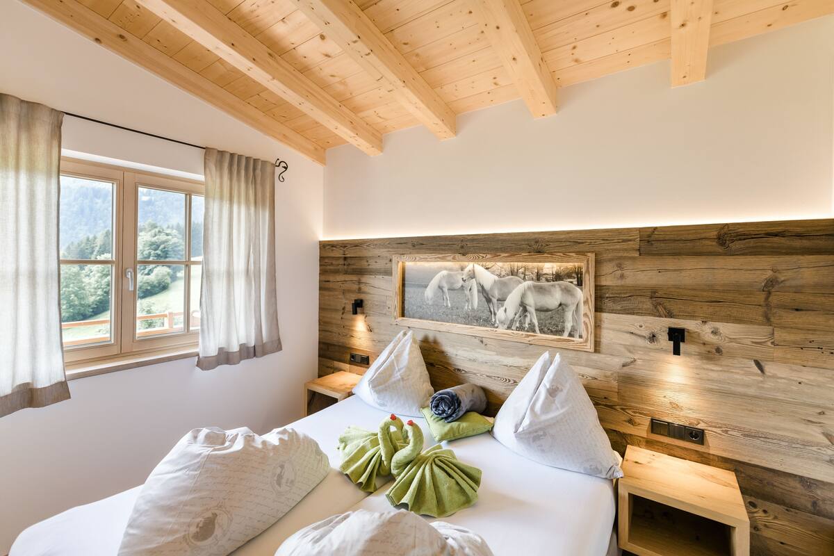 A bedroom with a wooden ceiling, a large window with curtains, a bed with pillows, a headboard with a picture, and bedside tables with lamps.