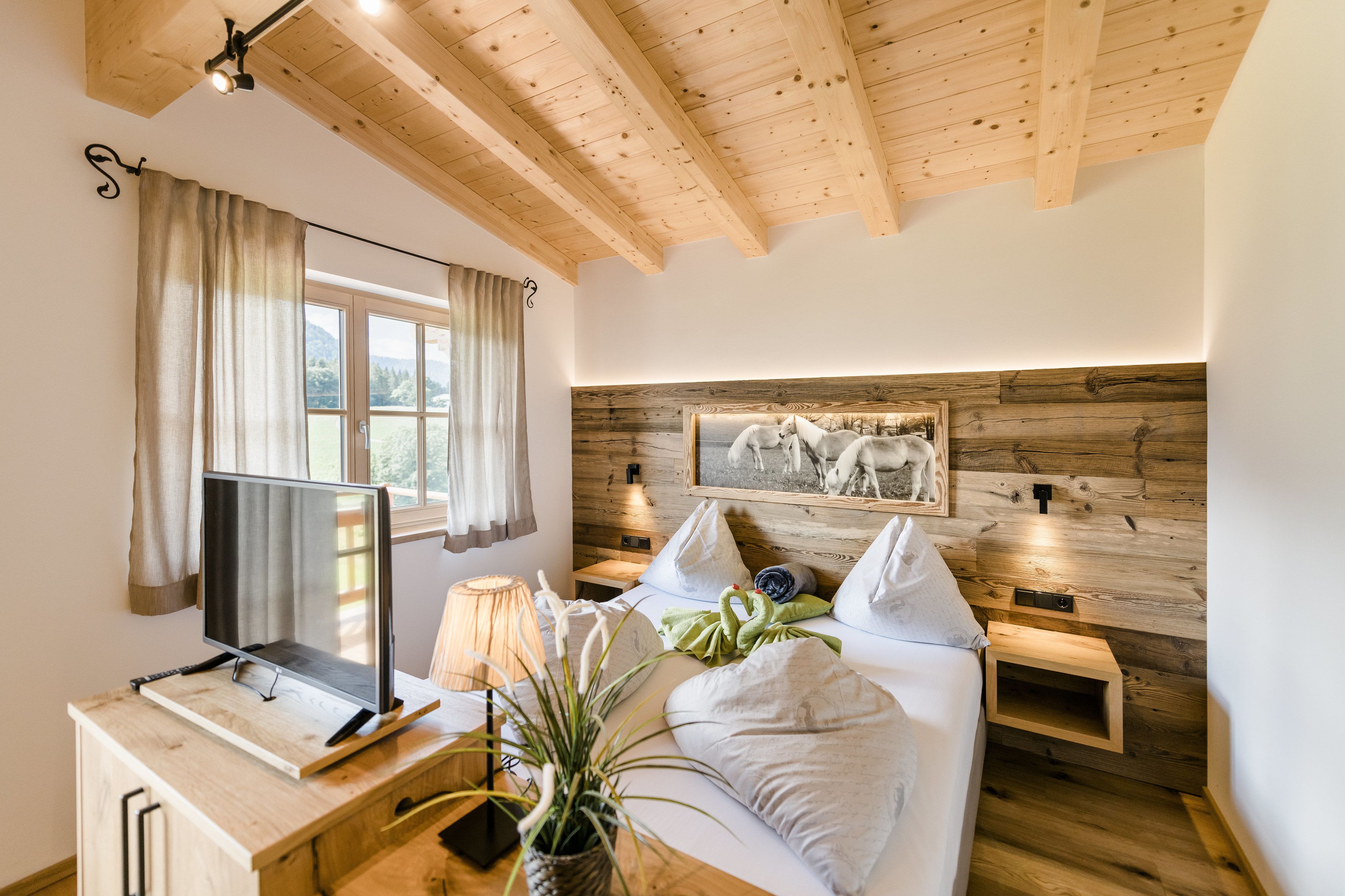 A bedroom with a wooden ceiling, a bed with white bedding, a mounted TV, a bedside table with a lamp, and a wall-mounted shelf.