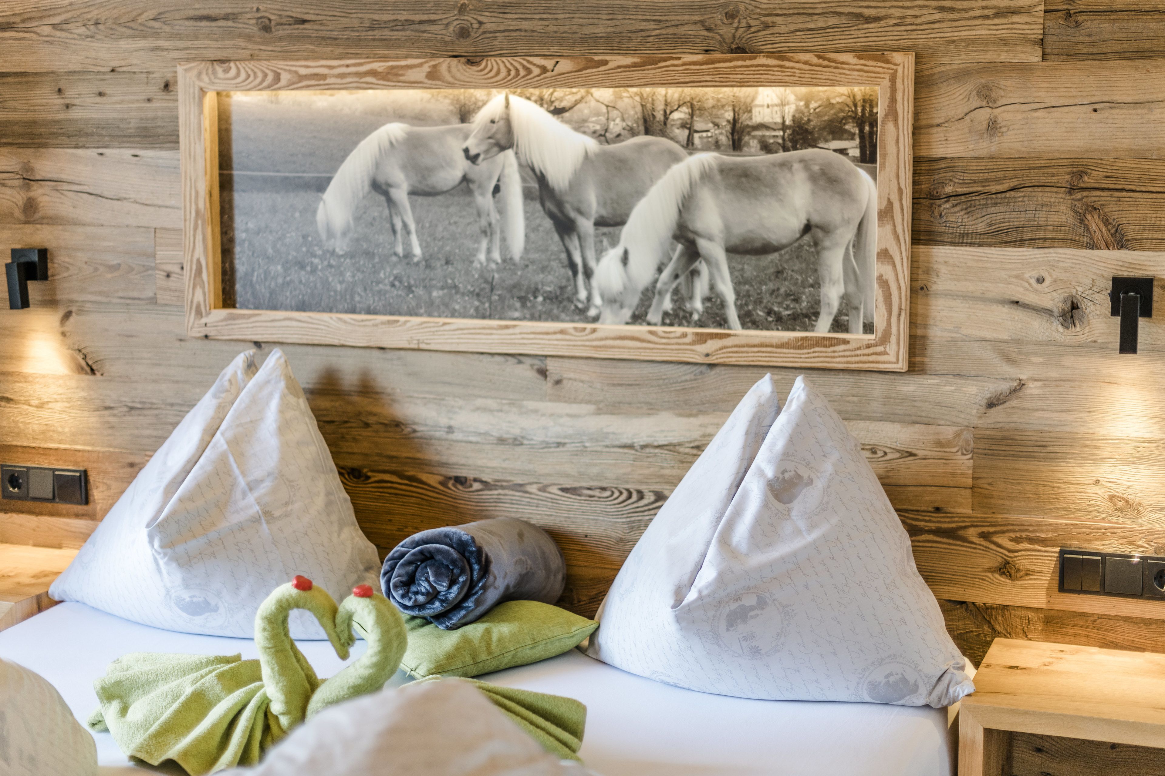 Pared de madera con fotografía en blanco y negro de un caballo enmarcado. Hay dos almohadas blancas y una toalla verde sobre una cama.