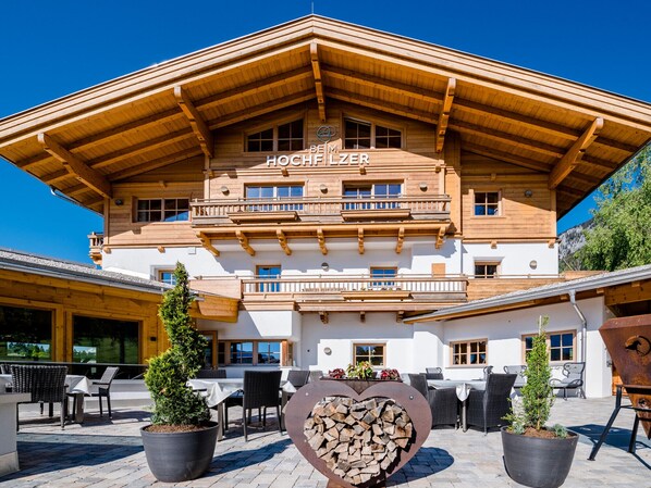 A wooden chalet with a sign reading "Hochfeler". It features outdoor seating with tables and chairs, potted plants, and a heart-shaped firewood display. - Beim Hochfilzer (Soell)