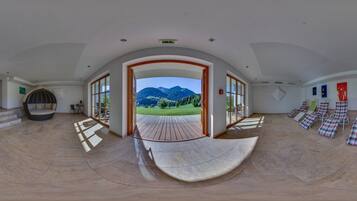 A panoramic view of a spacious room with a curved floor, a large window offering a mountain view, and several lounge chairs arranged along the window.