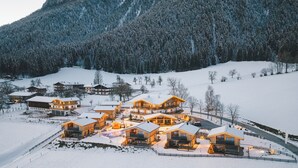 A snow-covered mountain landscape with a cluster of wooden houses with large windows and balconies, surrounded by snow-covered trees and a clear sky. - Beim Hochfilzer (Soell)
