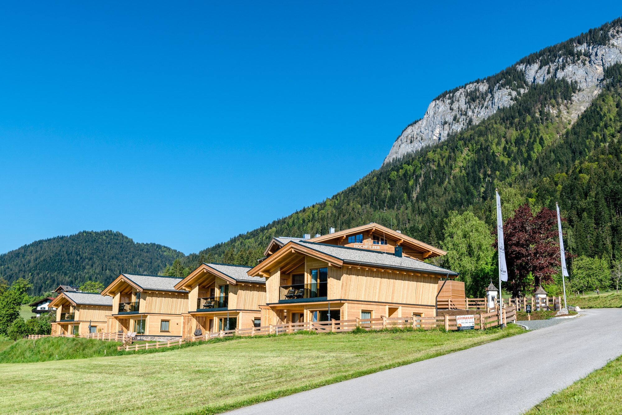 A wooden chalet-style hotel with multiple guest rooms, a mountain backdrop, and a clear sky.