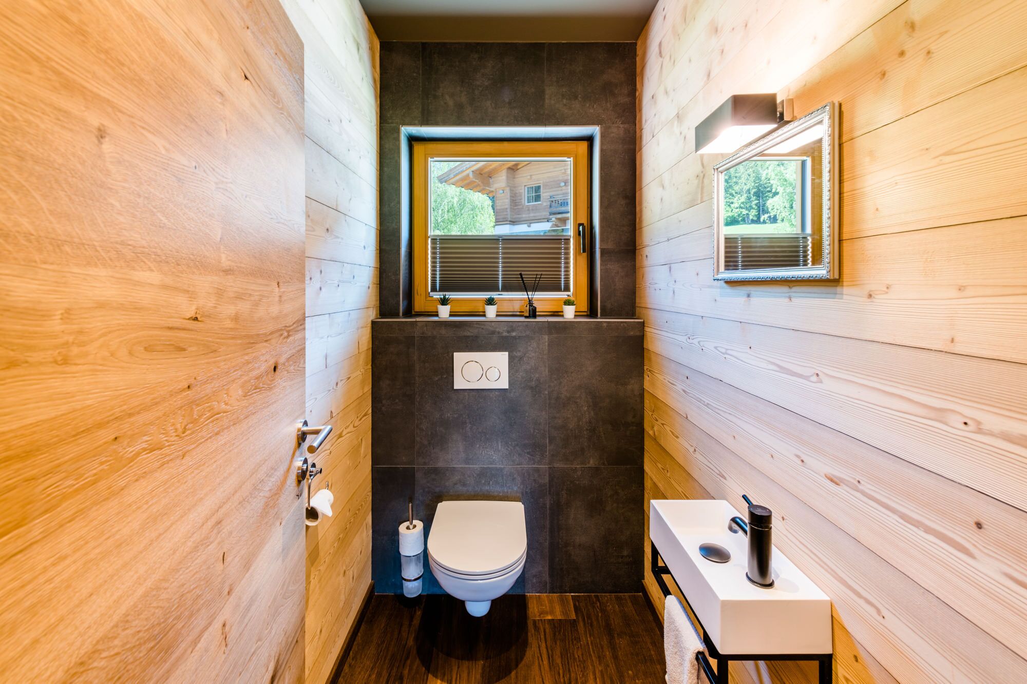 Une petite salle de bain aux murs en bois, avec des toilettes, un lavabo agrémenté d’une serviette et une fenêtre équipée de stores.