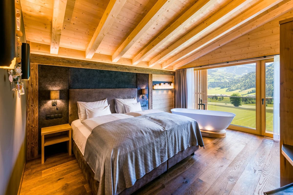 A bedroom with a large bed, a nightstand, a wall-mounted lamp, and a view of the outdoors through a large window.