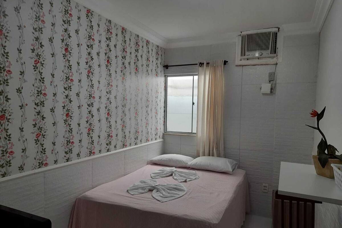 A bedroom with a bed, a window with curtains, a radiator, and a small table with a vase.