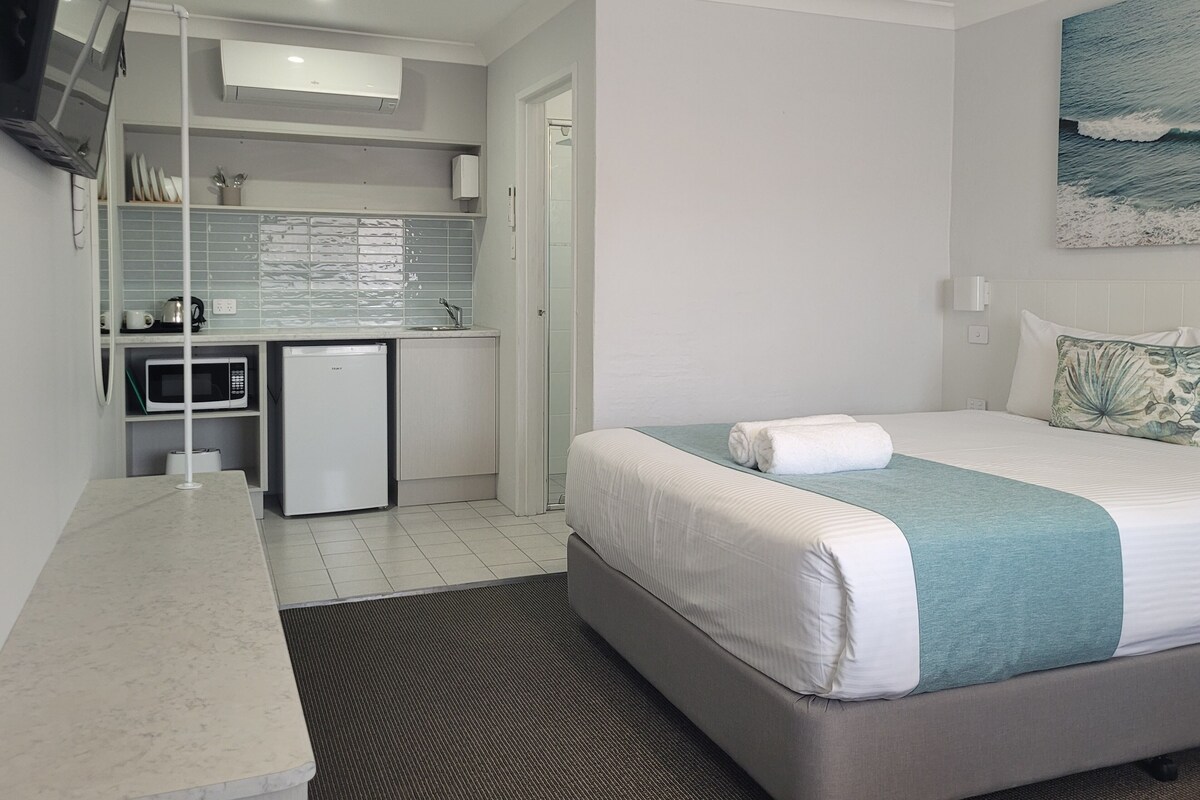 A compact hotel room with a bed, a small kitchenette, a TV mounted on the wall, and a painting of the ocean.