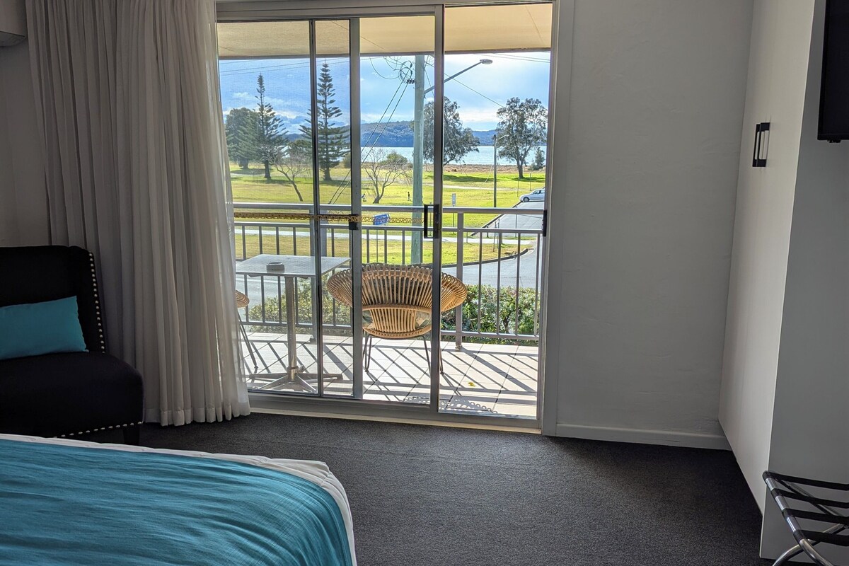 A hotel room with a bed, a chair, a balcony with a view of the outdoors, and a television on the wall.
