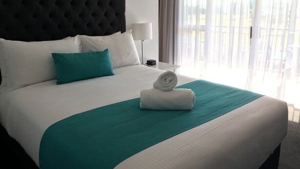 A neatly made bed with white and teal bedding, a black tufted headboard, and a rolled white towel.