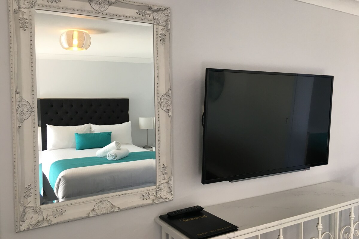 A hotel room with a bed, a television, a mirror, and a bedside table with a remote control and a guest information booklet.