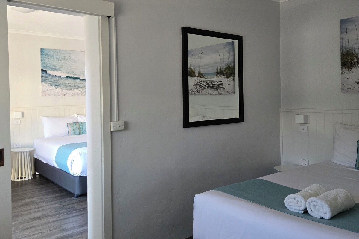 A hotel room with a bed, a small table, a chair, and two framed pictures on the wall.