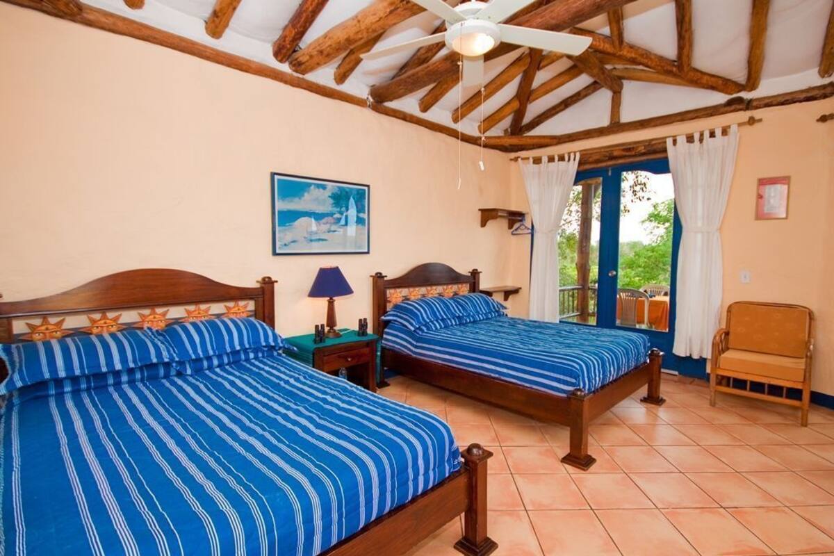 Ein Zimmer mit zwei Holzbetten, einem Deckenventilator, einem Gemälde, einem Vorhangfenster und einem Stuhl.