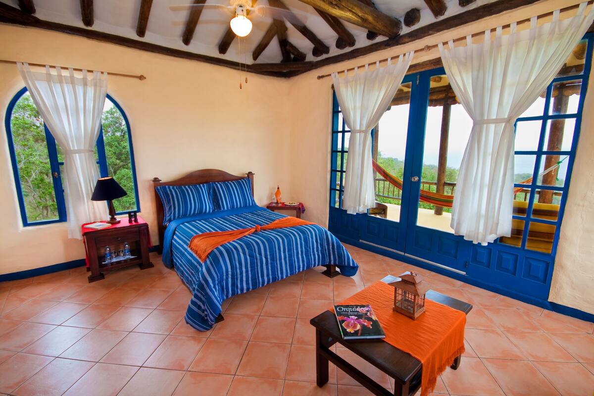 Ein Schlafzimmer mit Bett, Nachttisch, Tisch und Blick auf ein blaues Gebäude sowie Grünfläche.